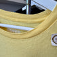 A close-up of a pale yellow crewneck t-shirt hanging on a white hanger, showing the ribbed collar, a white inner neck label reading 'SUPER', and a small sticker brand tag on the right chest.
