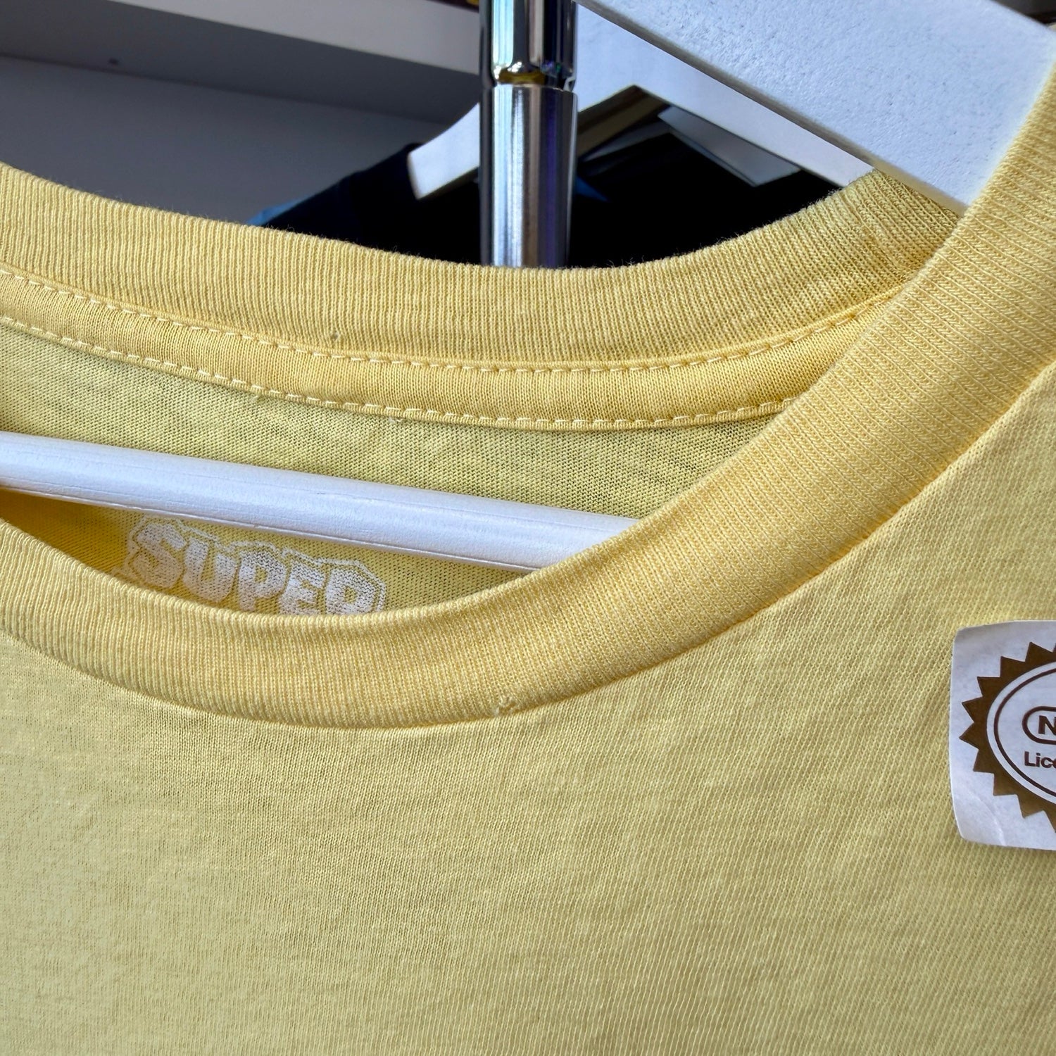 A close-up of a pale yellow crewneck t-shirt hanging on a white hanger, showing the ribbed collar, a white inner neck label reading 'SUPER', and a small sticker brand tag on the right chest.