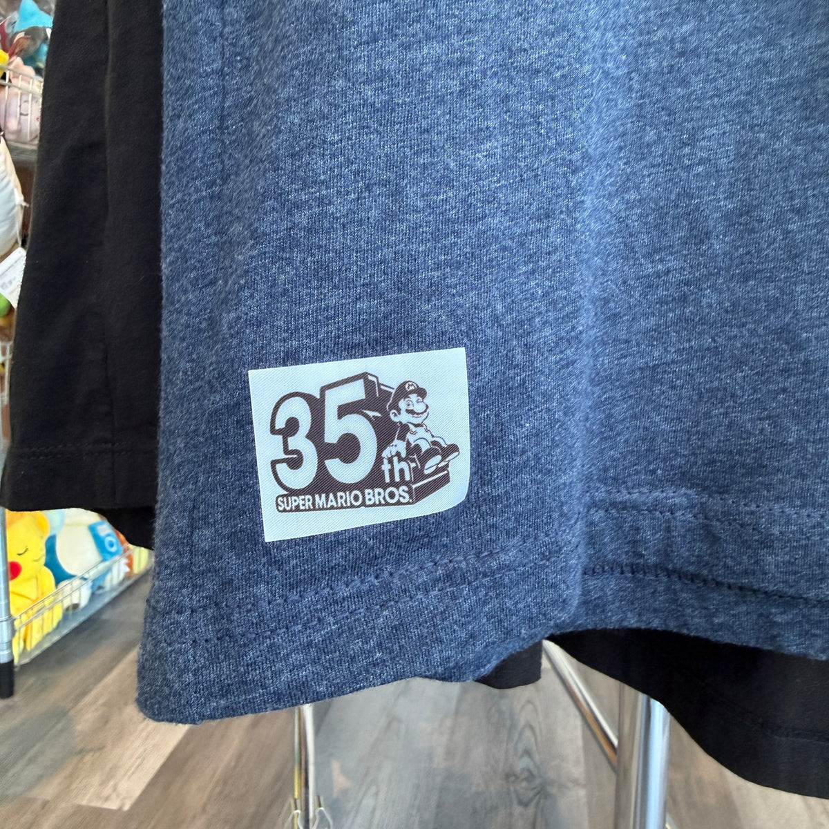 Close-up of a heathered blue t-shirt hanging on a rack, featuring a white rectangular patch on the sleeve with '35th Super Mario Bros.' and a cartoon character graphic.