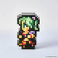 A pixel-art character figurine made of black square blocks, with green hair, yellow and orange clothing, and small pink and purple accents, photographed on a clean light gray background. A small '© SQUARE ENIX' mark appears in the bottom right corner.