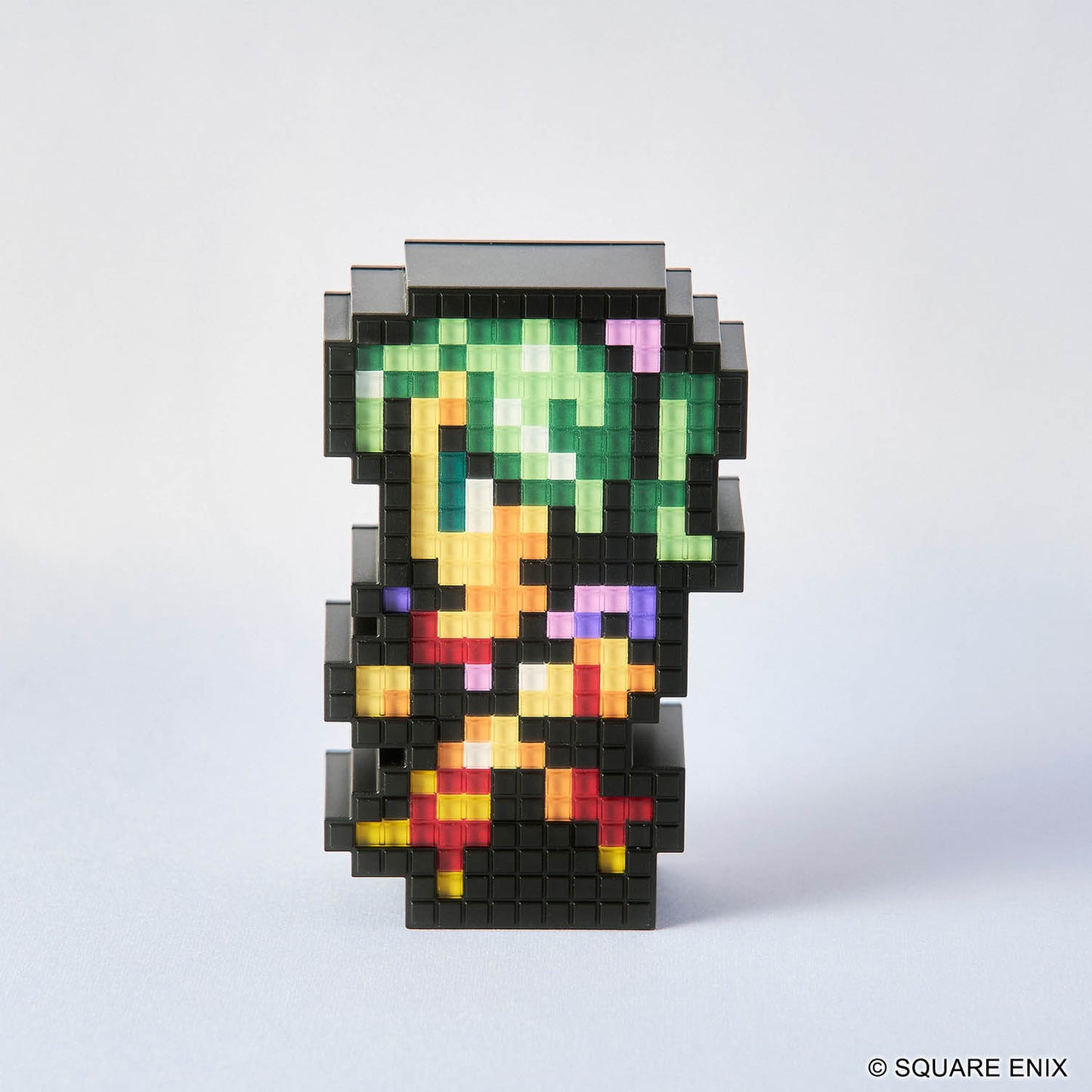 A pixel-art character figurine made of black square blocks, with green hair, yellow and orange clothing, and small pink and purple accents, photographed on a clean light gray background. A small '© SQUARE ENIX' mark appears in the bottom right corner.