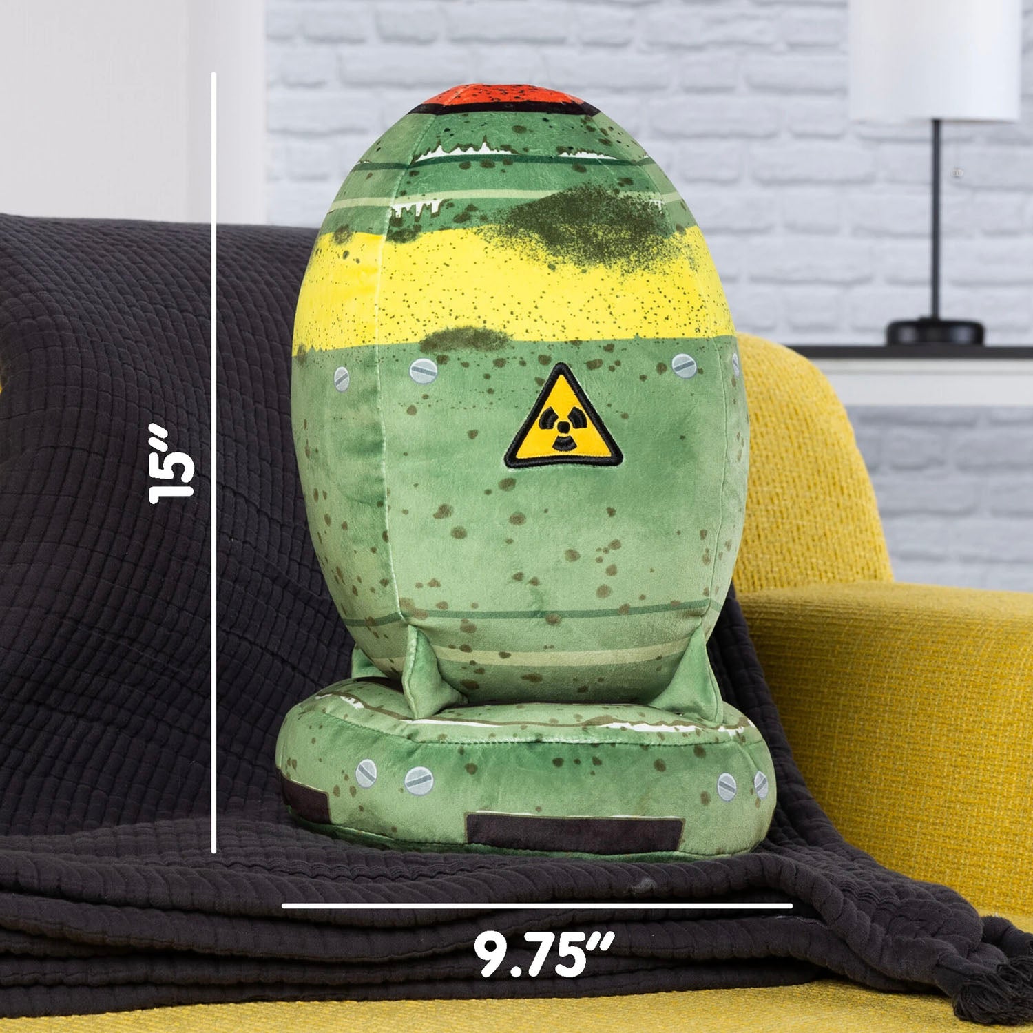 Nuke shaped plush toy on a black blanket laid across a yellow sofa. Living room in the background. White text and lines show measurements of 15 by 9.75 inches.
