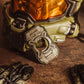 A close-up of a sci-fi helmet with an olive green body and reflective orange visor, alongside a matching metallic bottle opener and dark gloves on a textured brown surface.
