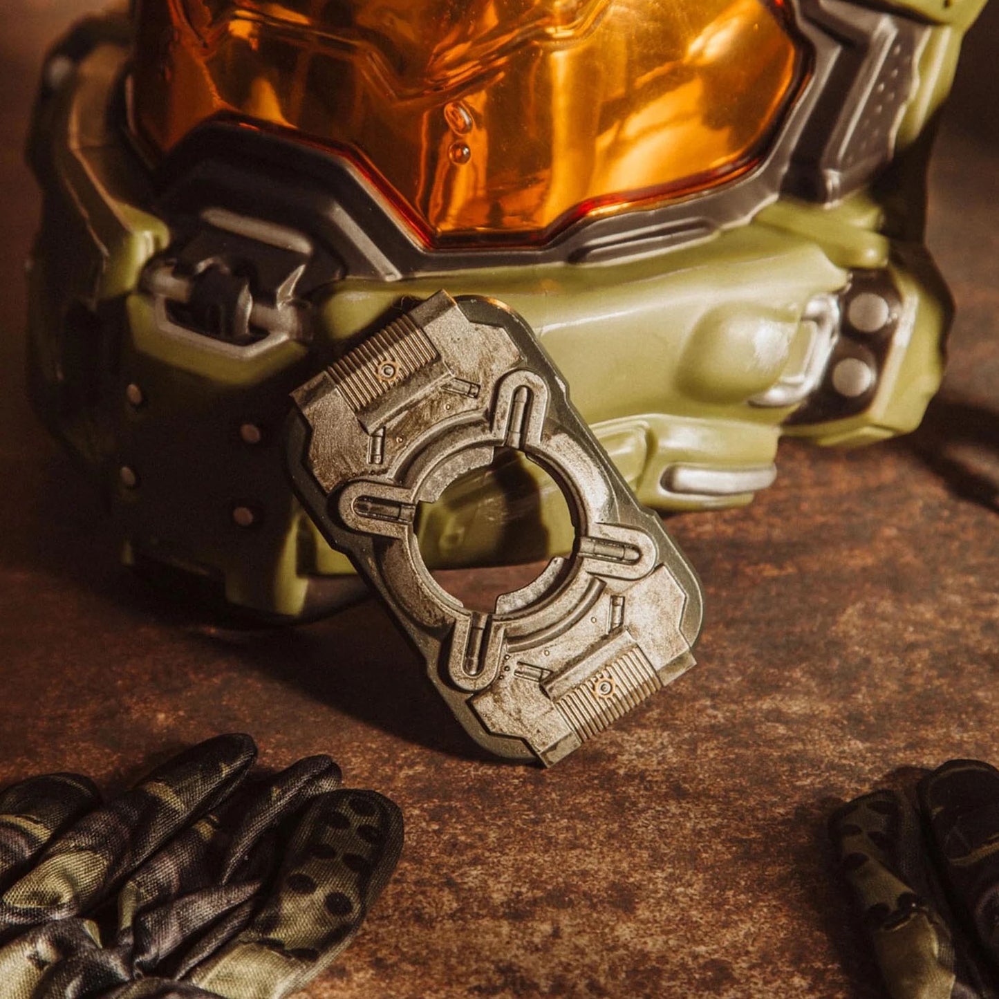 A close-up of a sci-fi helmet with an olive green body and reflective orange visor, alongside a matching metallic bottle opener and dark gloves on a textured brown surface.