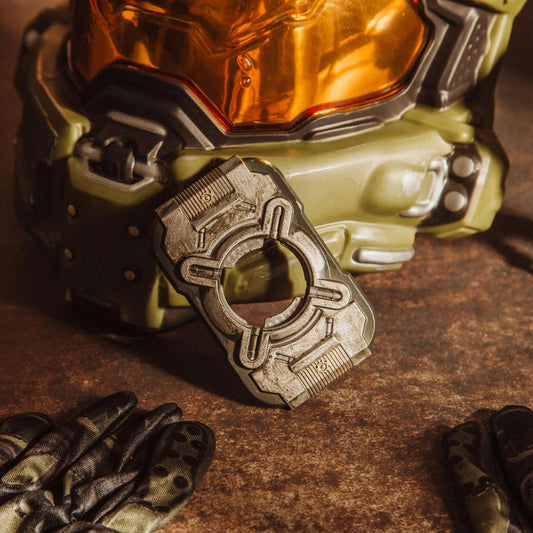 A close-up of a sci-fi helmet with an olive green body and reflective orange visor, alongside a matching metallic bottle opener and dark gloves on a textured brown surface.