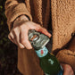 A close-up of a person's hand using a silver metal bottle opener to open a green glass bottle, wearing a brown textured fleece jacket.
