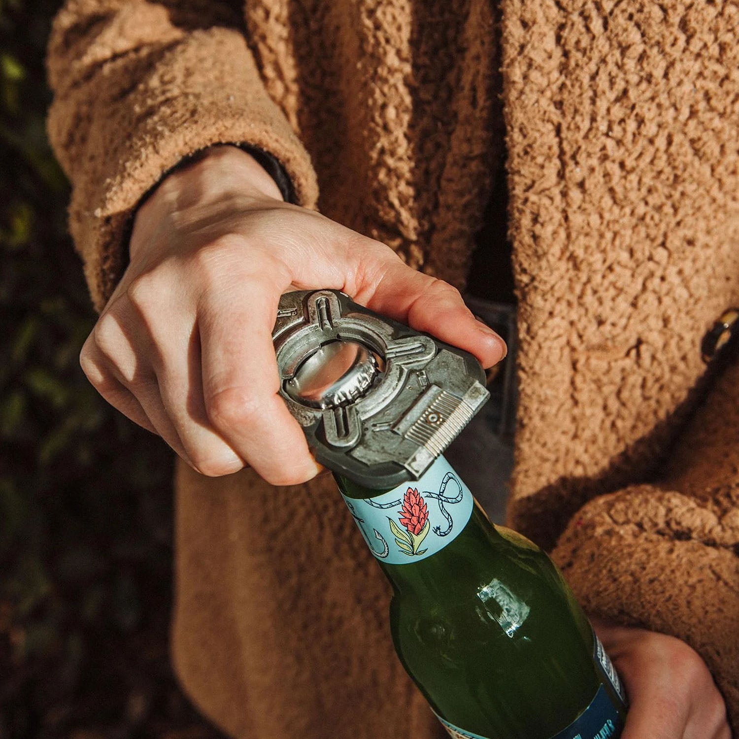 A close-up of a person's hand using a silver metal bottle opener to open a green glass bottle, wearing a brown textured fleece jacket.