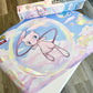 A Pokémon-themed playmat featuring Mew floating in a bubble among pastel clouds, with a matching Pokémon playmat box partially visible behind it on a wooden floor.
