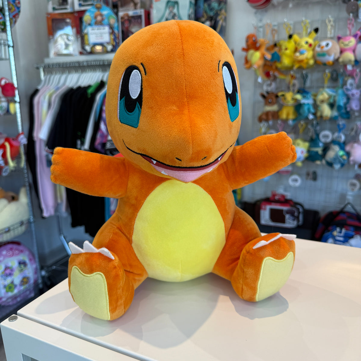 A Charmander plush toy sitting on a white table in a retail store. The plush is orange with a yellow belly and blue eyes, with its arms outstretched.