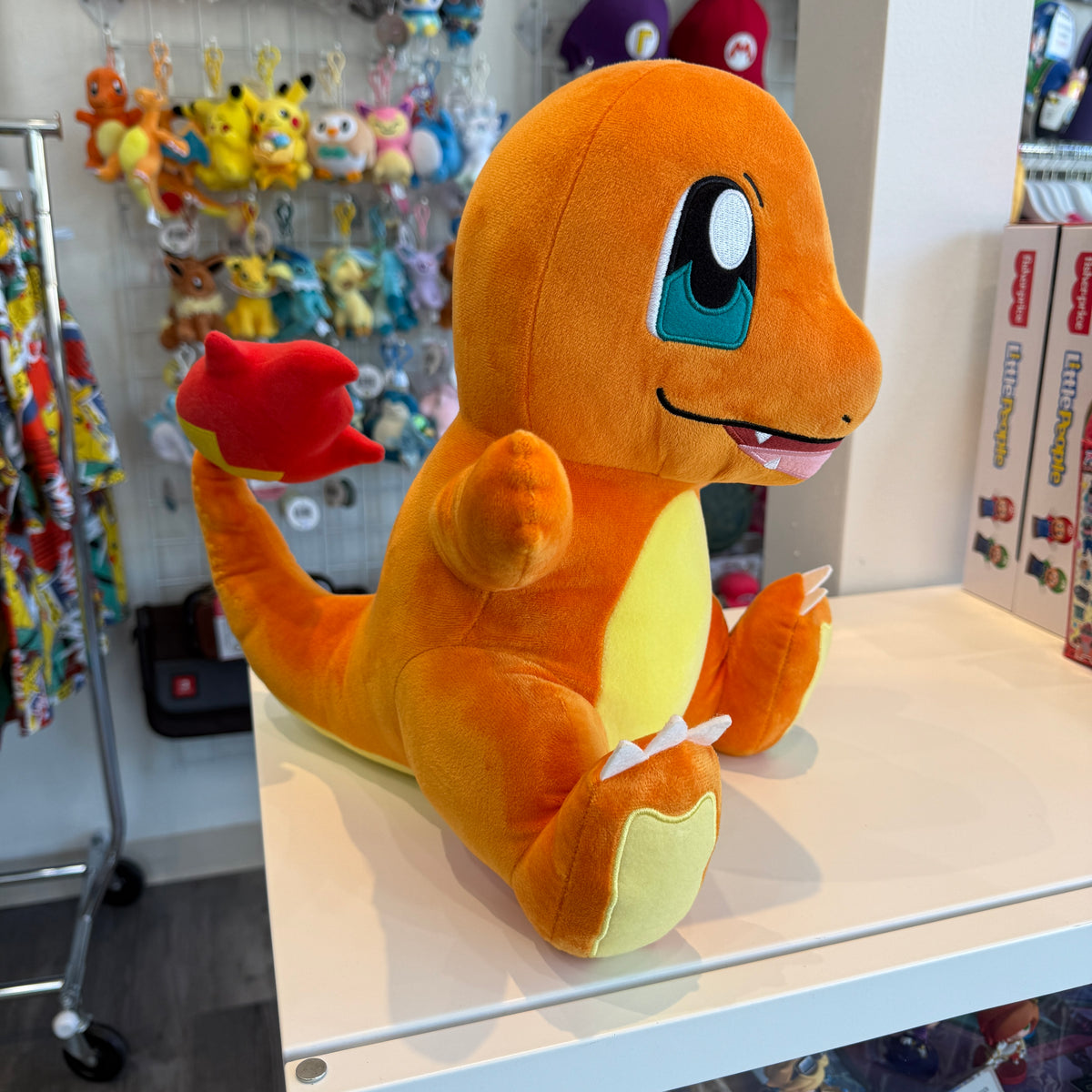 A Charmander plush toy sitting on a white table in a retail store. The plush is orange with a yellow belly and blue eyes, with its arms outstretched.