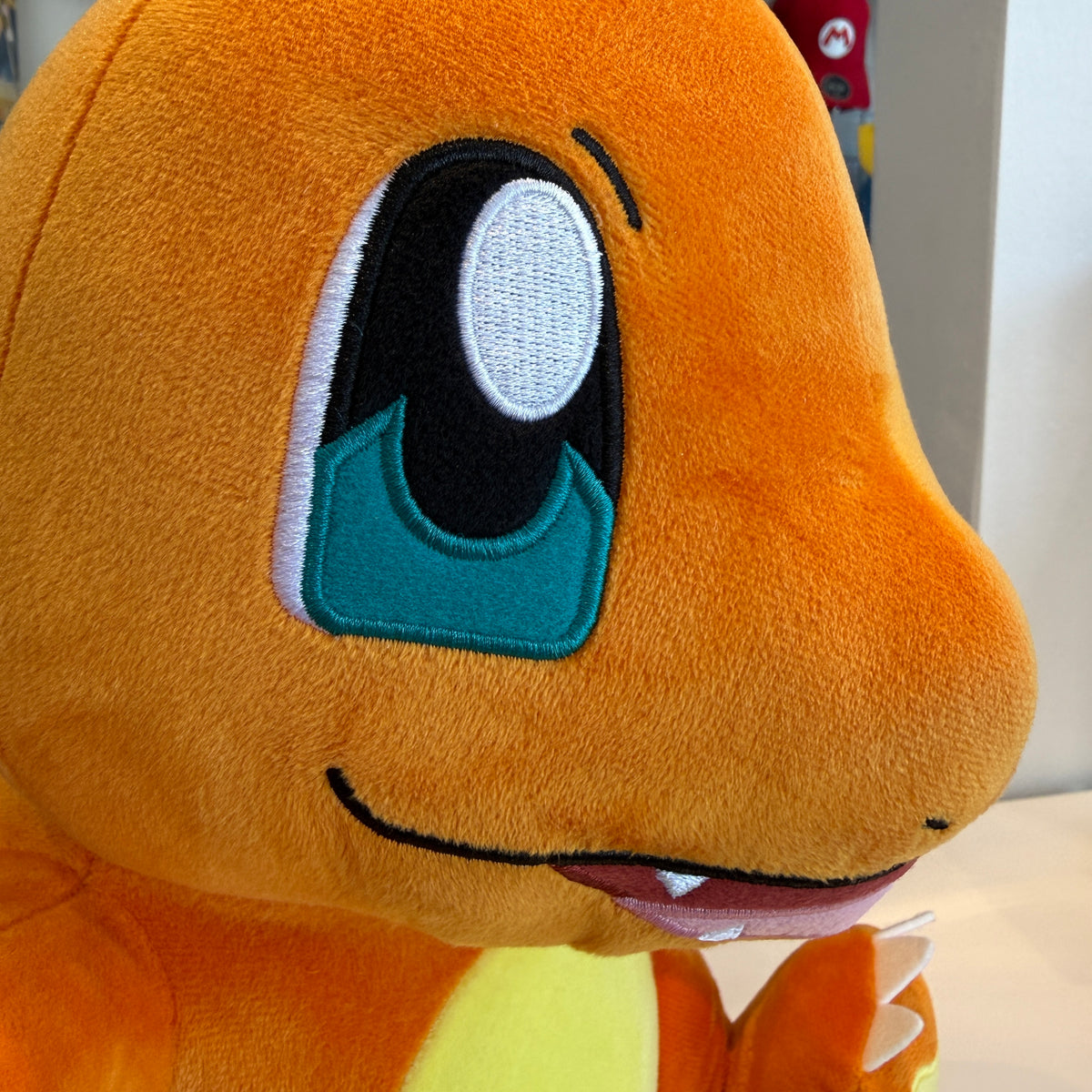 Close up of a  Charmander plush toy sitting on a white table in a retail store. The plush is orange with blue and white embroidered eyes.