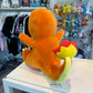 Backview of a Charmander plush toy sitting on a white table in a retail store. The plush is orange with a yellow belly and blue eyes, with its arms outstretched.