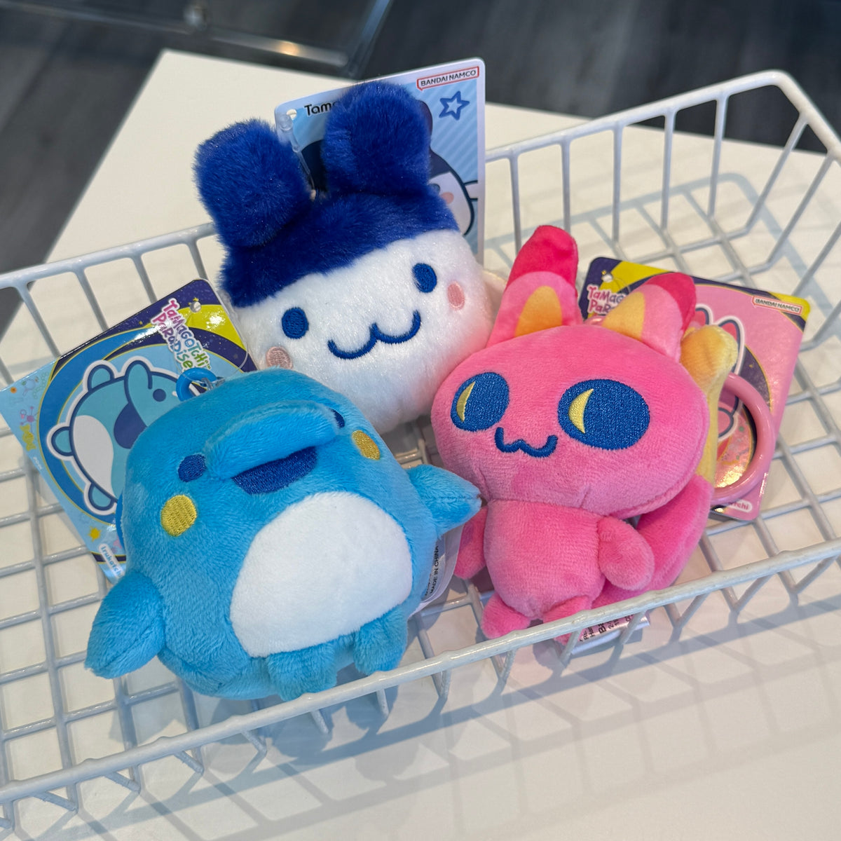 Three colorful Tamagotchi themed plush toys sitting in a white, wire mesh basket. 