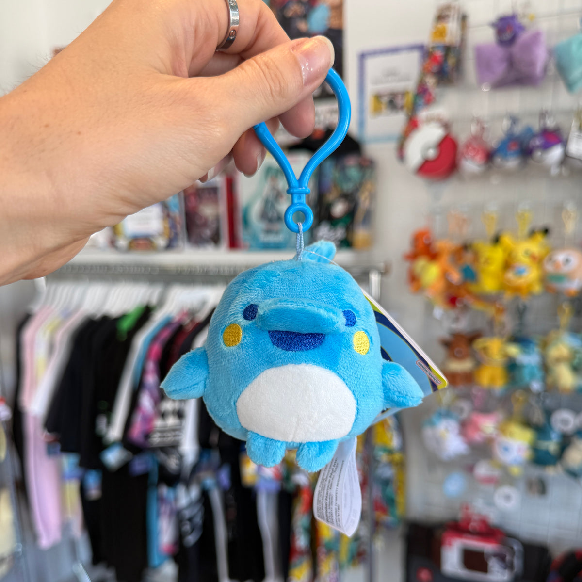 Hand holding blue dolphin Tamagotchi character with a store in the background. 