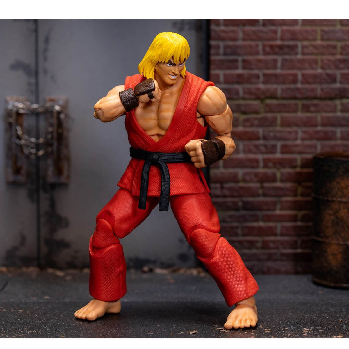 A Street Fighter Ken action figure posed in a fighting stance, wearing his signature red gi with a black belt and brown wrist wraps, set against a backdrop of a brick wall and dark alley floor.