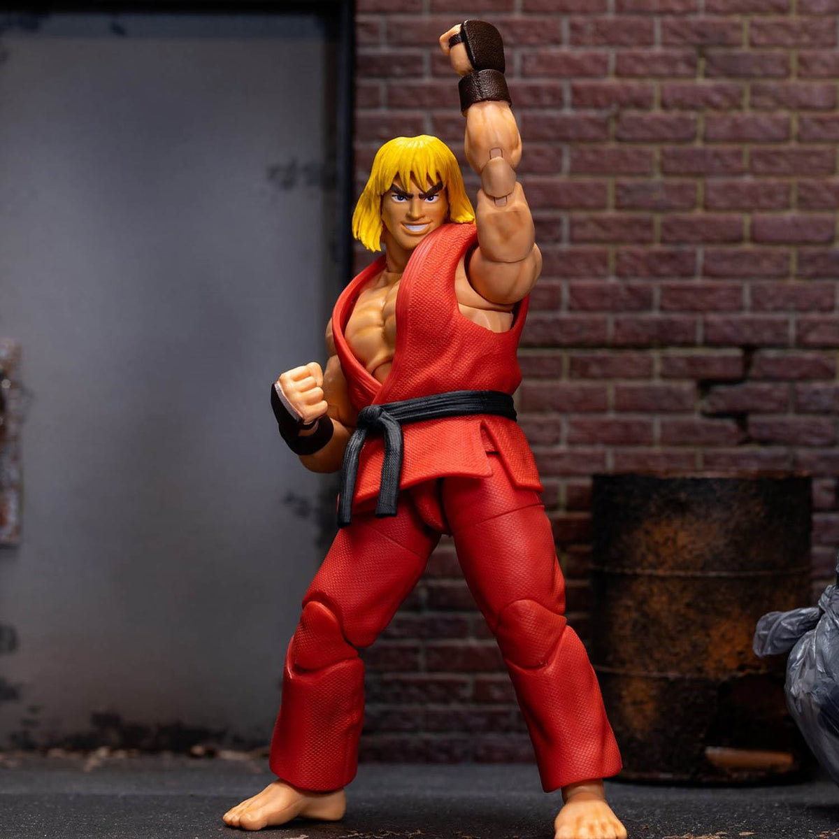 Street Fighter Ken action figure posed in a fighting stance, wearing a red gi with a black belt and brown finger wraps, set against a brick wall and barrel backdrop.