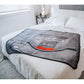 A gray plush blanket featuring a Sega Genesis 16-BIT graphic design, draped over a bed with white sheets in a bedroom setting.