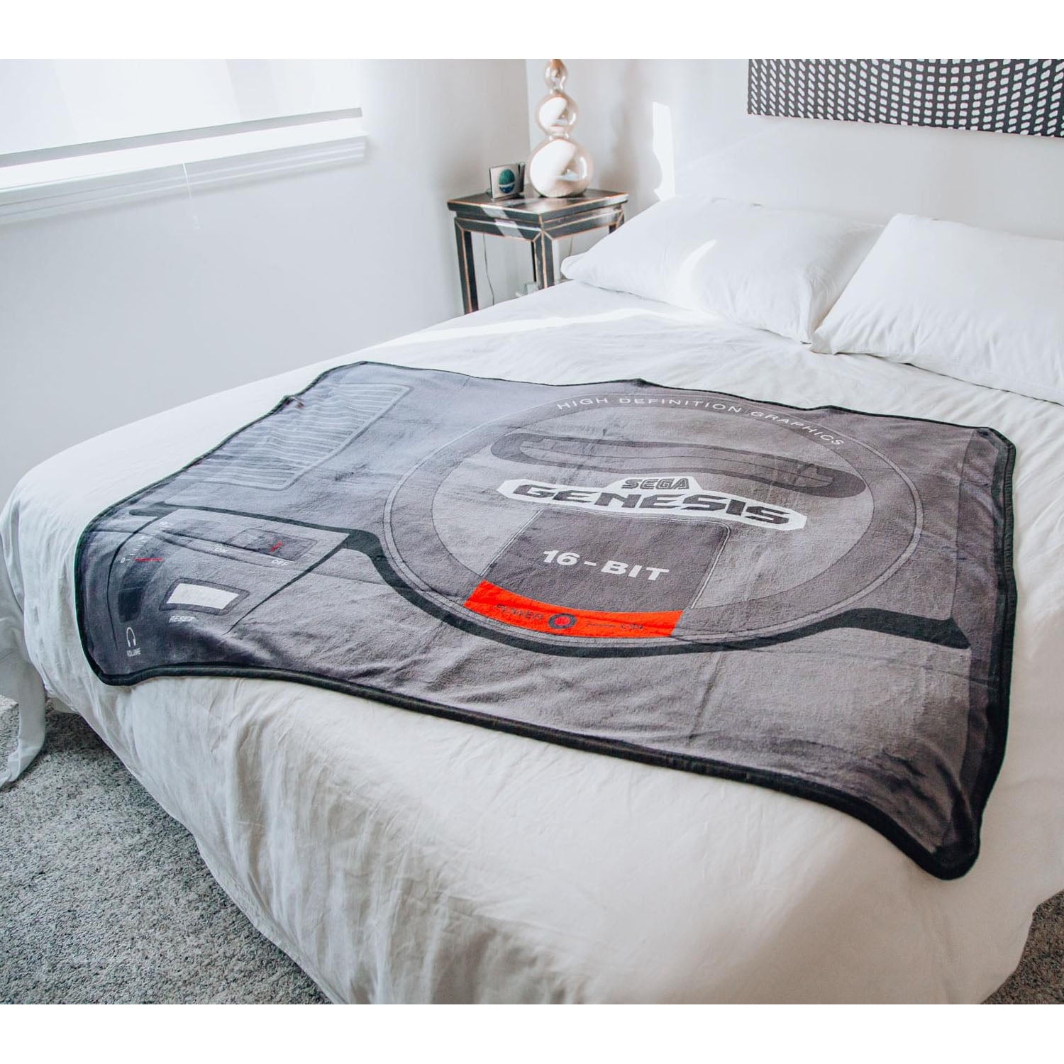 A gray plush blanket featuring a Sega Genesis 16-BIT graphic design, draped over a bed with white sheets in a bedroom setting.